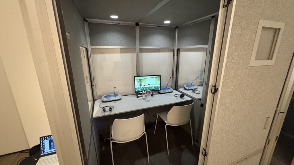 Tolkencabine met twee werkplekken, koptelefoons en een scherm met live beeld van de zaal